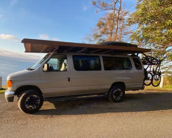 Tan adventure campervan with extended wooden roof, cargo box, and bikes parked by the ocean. Perfect for an RV rental trip.. Ford E350 Box Van 2003
