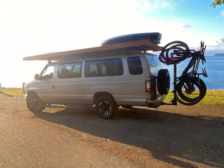 Adventure-ready RV rental van with roof rack, cargo box, and bike rack, parked by serene water for scenic trips.. Ford E350 Box Van 2003