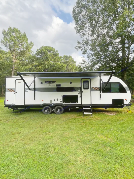 Spacious white travel trailer RV rental with awning extended, nestled in a grassy outdoor setting with trees.. Forest River Wildwood X-Lite Platinum 2024