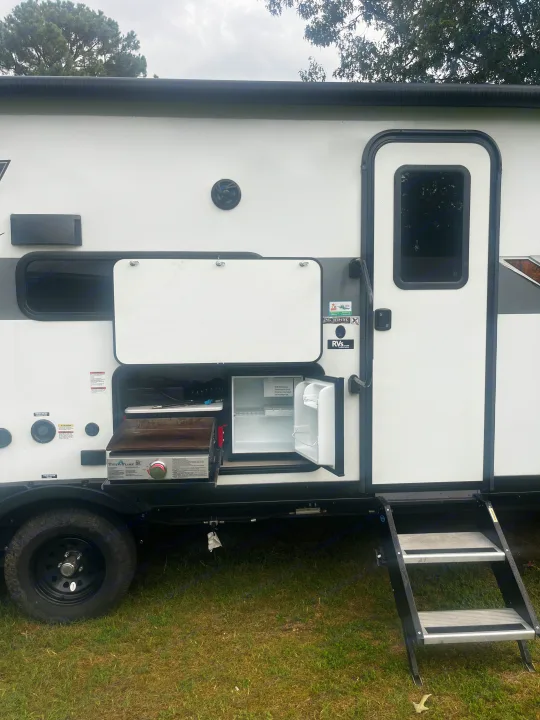 Side of an RV rental with its outdoor kitchen open, revealing a griddle and mini-fridge. Steps lead to the door.. Forest River Wildwood X-Lite Platinum 2024