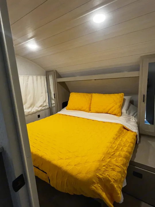 Bright yellow bed in a compact RV rental bedroom, featuring a plank ceiling, window curtains, and built-in storage.. Forest River Wildwood X-Lite Platinum 2024