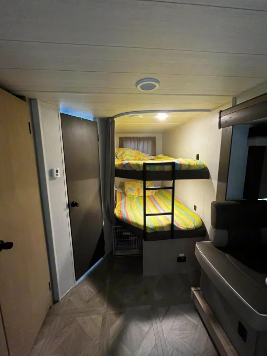 This RV rental features a cozy interior with bunk beds, colorful bedding, a pet crate, and a small seating area.. Forest River Wildwood X-Lite Platinum 2024
