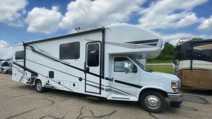 Side view of a white Class C RV rental with dark windows and grey/black graphics, parked on asphalt under a cloudy sky.. Jayco Greyhawk 2026