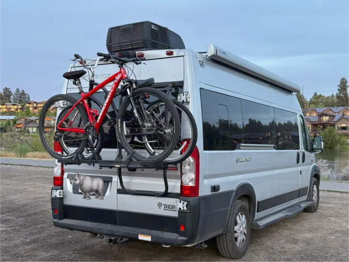 Silver camper van RV rental with two bikes on a rear rack, parked by a lake. Ready for adventure!. Thor Motor Coach Sequence 2022
