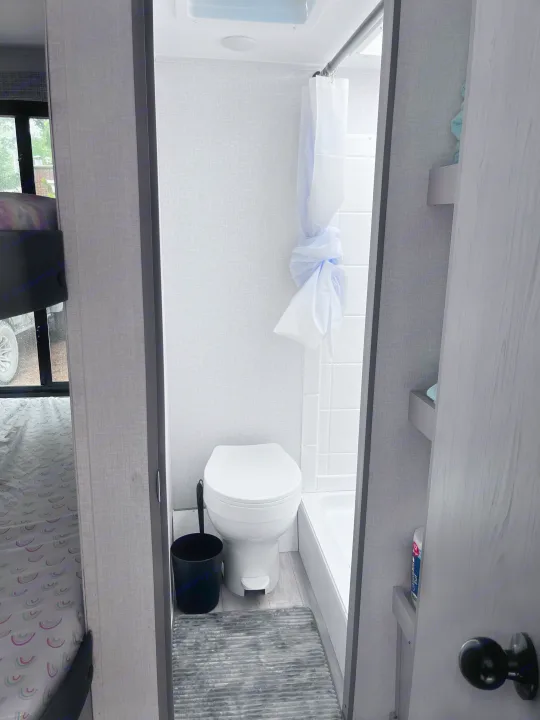 Compact RV rental bathroom: features a white toilet, shower, and grey rug. Bunk beds seen to the left.. Clipper 4K 2023