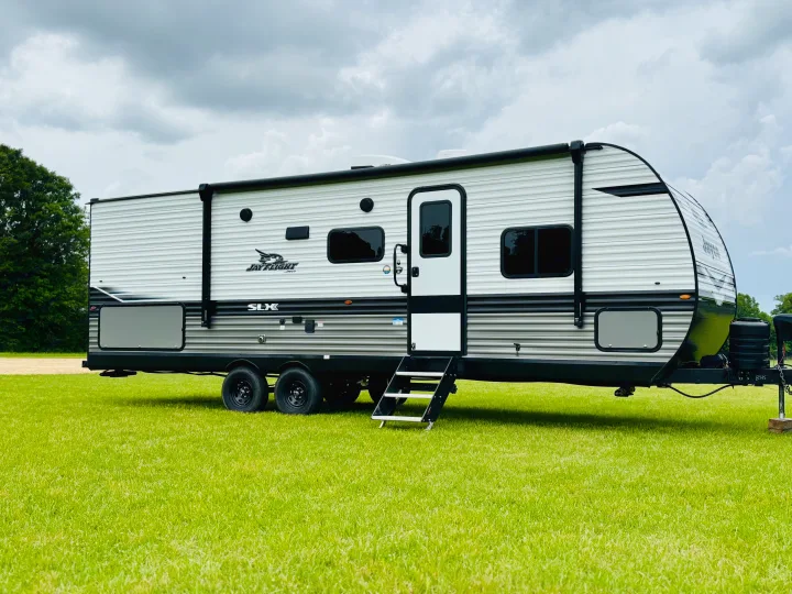This modern white and gray travel trailer, great for RV rental, sits on bright green grass under a cloudy sky. Ideal for camping.. Jayco Jay Flight SLX 2024