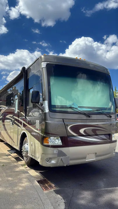 A grey Thor Palazzo RV rental, parked on a sunny street with a blue sky, shows damage on its front bumper.. Thor Palazzo 2014