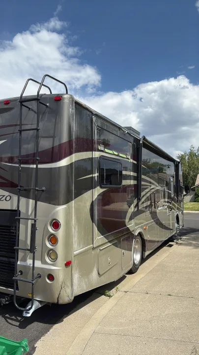 A dirty, two-tone gray and maroon RV rental, viewed from its rear passenger side, parked on a roadside under a cloudy sky.. Thor Palazzo 2014
