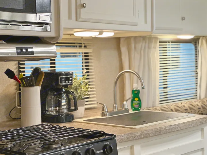 RV rental kitchen: compact stove, microwave, sink with cover, coffee maker, and cooking utensils, well-lit by under-cabinet lights.. Coachmen Apex LE 2017