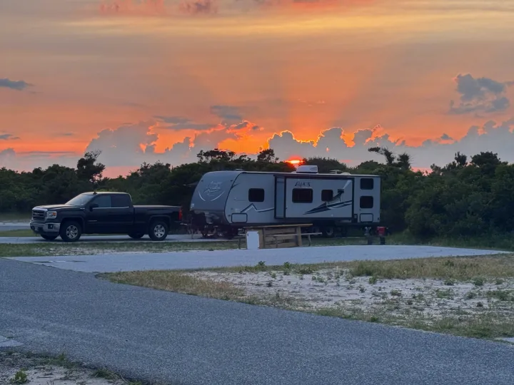 A grey travel trailer (RV rental) and black pickup truck are parked at a gravel campsite during a vibrant sunset.. Coachmen Apex LE 2017