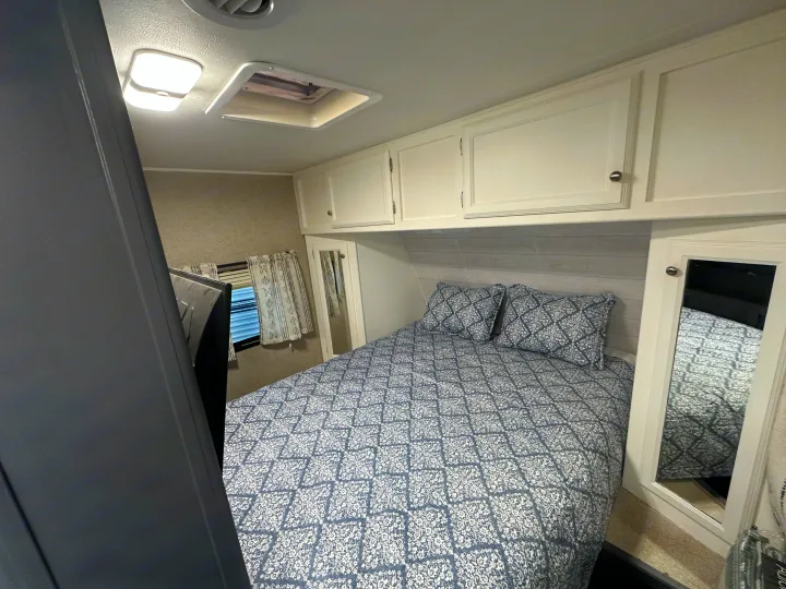 RV rental bedroom with blue/white patterned bedding, white overhead cabinets, and a window, offering a cozy retreat.. Coachmen Apex LE 2017