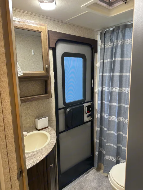 This RV rental features a practical bathroom with a sink, mirror cabinet, door, shower curtain, and toilet.. Coachmen Apex LE 2017