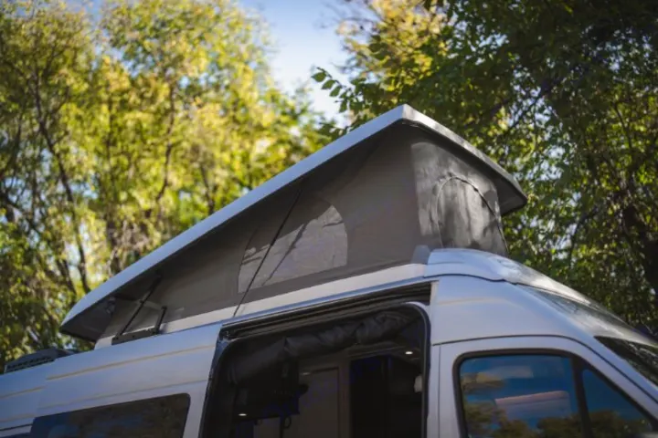 Pop-top camper van with extended roof & open side door, showcasing its versatile design for an RV rental. Amidst green trees.. Mercedes Sprinter 2023