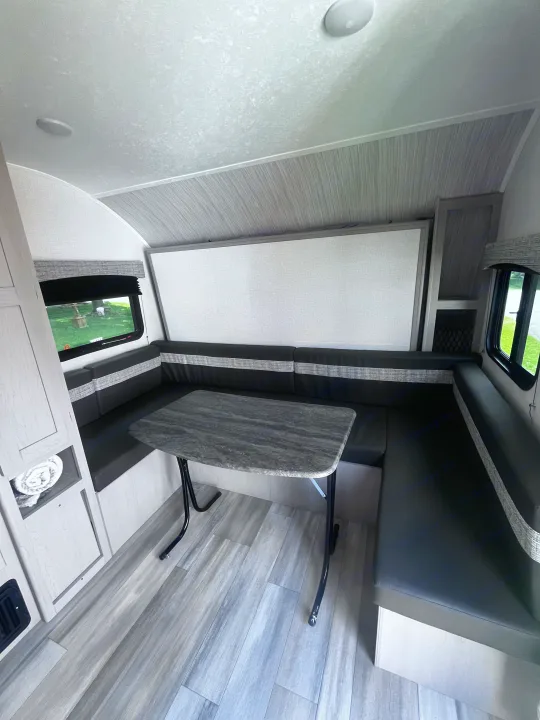 RV rental interior: A U-shaped dinette with dark cushions, gray table, and light wood flooring, with windows showing outside.. Clipper 4K 2023