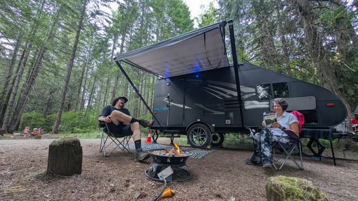 A couple relaxes by a gas campfire with their RV rental in a serene forest campground. One person reads, the other reclines.. Travel Lite Falcon 2018