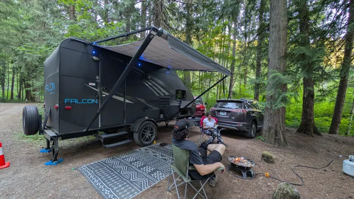 Enjoying a peaceful evening at a forest campsite with an rv rental, featuring a Falcon travel trailer, fire pit, and people relaxing outdoors.. Travel Lite Falcon 2018