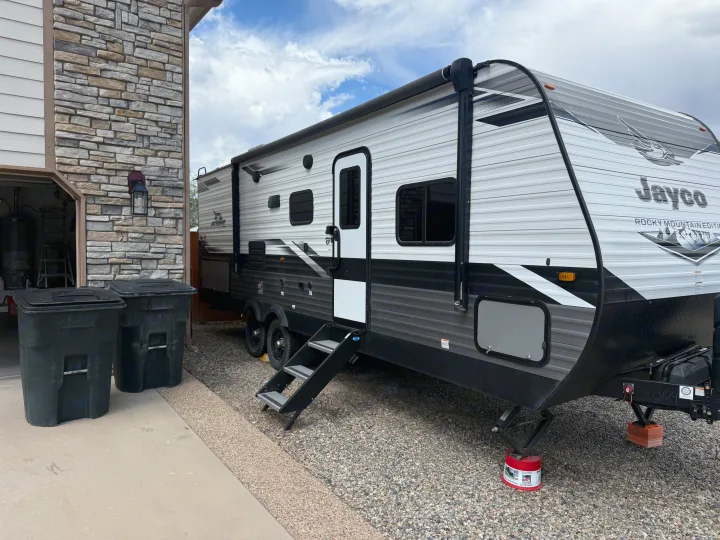 Modern grey and white Jayco travel trailer, suitable for an rv rental, parked on gravel next to a stone house under cloudy skies.. Jayco Jayco Rocky Mountain 2022