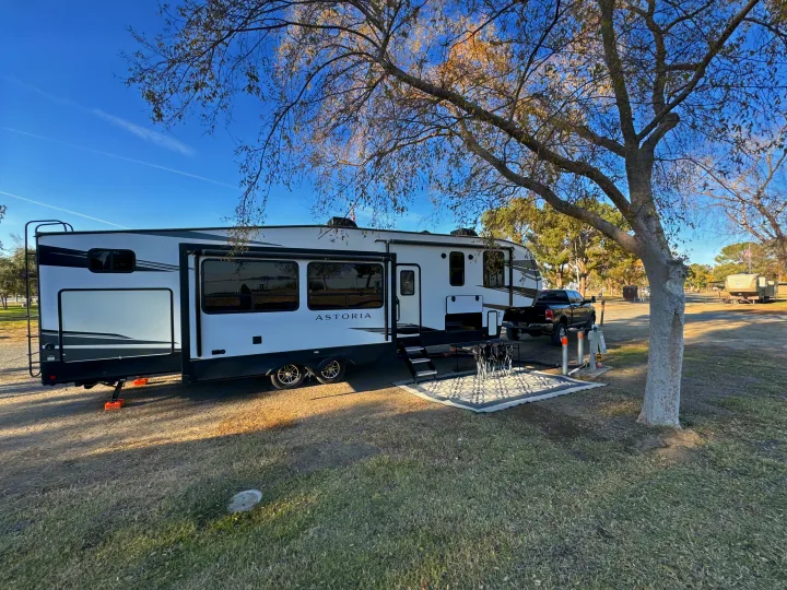 RV rental: White and black fifth-wheel RV and truck at a sunny campsite with an outdoor rug and chairs.. Astoria 1500 2021