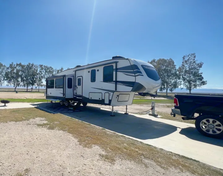 A large white Astoria fifth-wheel RV rental parked at a sunny campsite with a truck, perfect for a peaceful outdoor escape.. Astoria 1500 2021