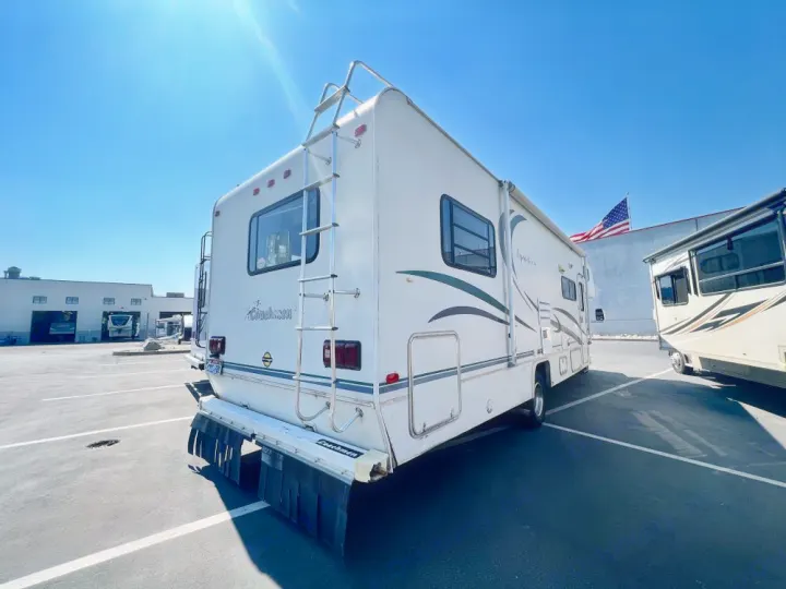 A white Coachmen RV rental, seen from the rear, is parked in a sunny lot with other RVs, featuring a rear ladder.. Coachmen Leprechaun 2001