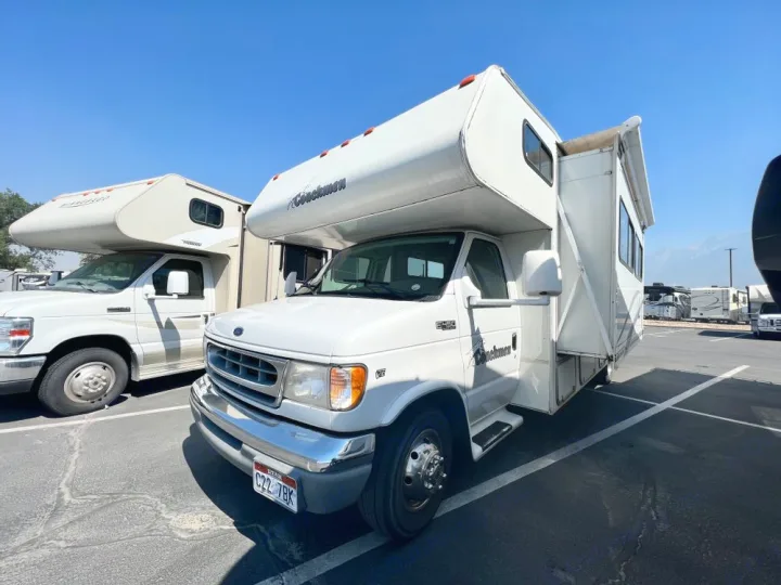 Two white Class C motorhomes are parked in a lot under a clear sky. The front Coachmen RV with a slide-out is perfect for rv rental.. Coachmen Leprechaun 2001