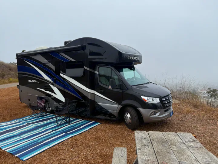 Black Tiburon SV RV rental set up with outdoor seating and striped mat at a misty viewpoint.. Thor Tiburon 2022