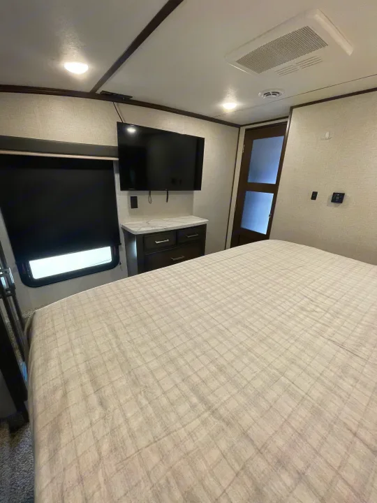 Step inside this cozy RV rental bedroom, featuring a large bed, wall-mounted TV, dresser, and a door for privacy.. Dutchmen 3602LFKB 2021