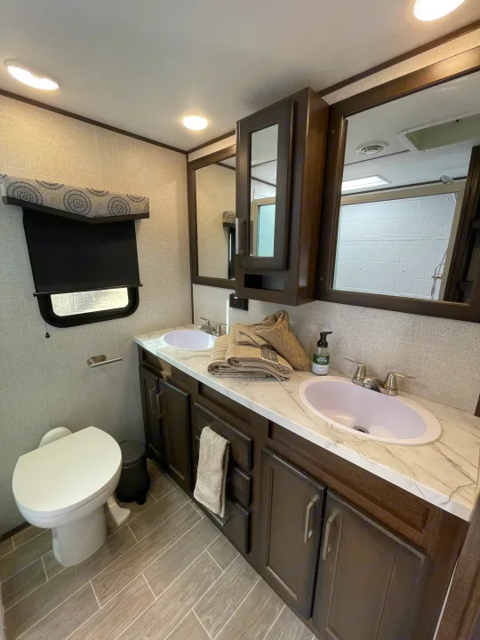 Bright, modern RV rental bathroom: dual sinks, dark vanity, toilet, window, and wood-look floor.. Dutchmen 3602LFKB 2021