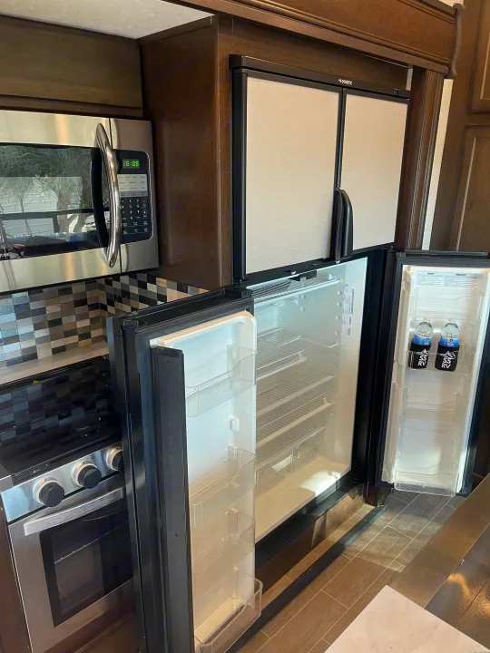 An rv rental kitchen with stainless steel microwave, stove, and open Dometic fridge displaying empty shelves and water bottles.. Dutchmen 3602LFKB 2021