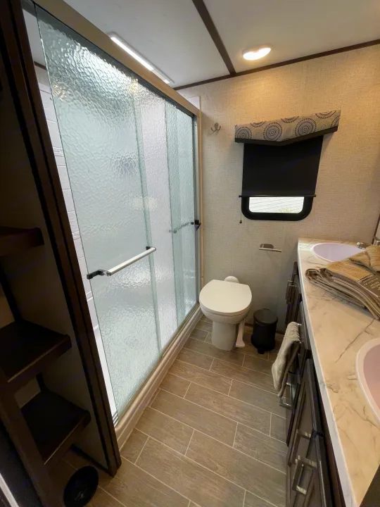 Bright RV rental bathroom with a glass shower, toilet, and dual-sink vanity, featuring wood-look tile flooring.. Dutchmen 3602LFKB 2021