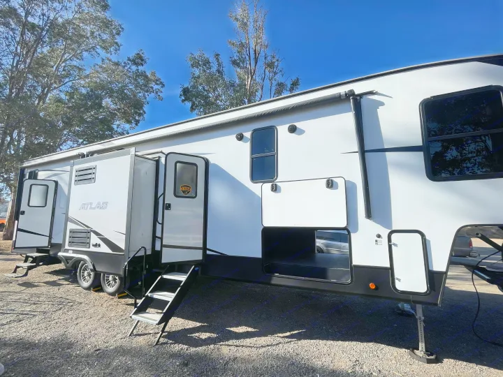 A large white Atlas brand RV rental set up outdoors on gravel with steps and storage open, under a clear blue sky.. Dutchmen 3602LFKB 2021