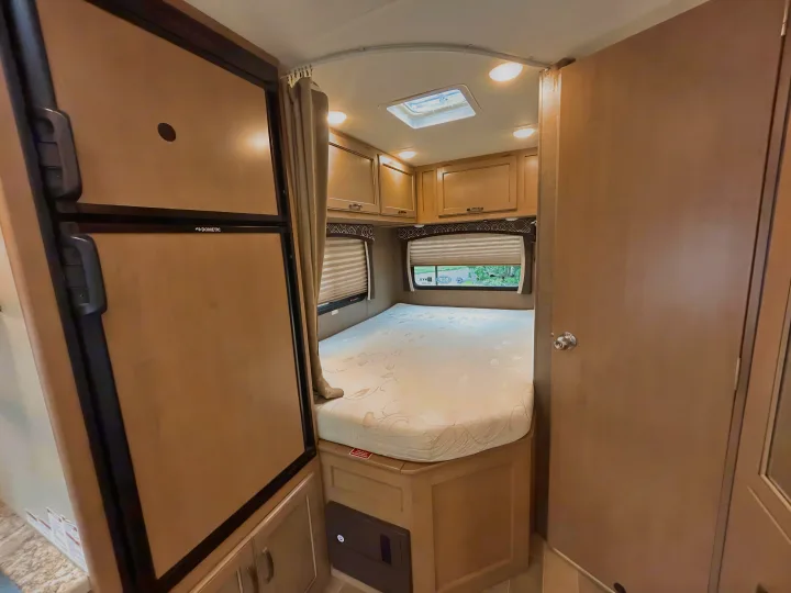 This RV rental interior shows the bedroom with a bed, windows, and overhead cabinets, beside a Dometic refrigerator.. Thor Chateau 2019