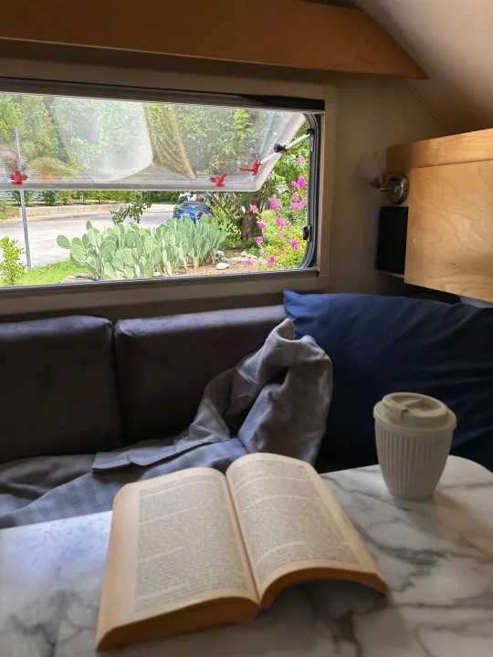 Cozy RV rental: a book and coffee cup on a table, with an open window view of prickly pear cacti & greenery outside.. nuCamp T@B 2022