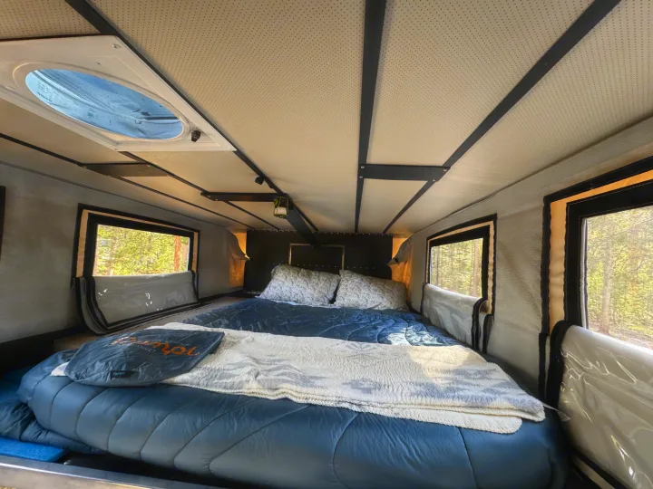 Inside a comfortable RV rental bedroom, featuring a large bed, side windows with tree views, and a Dometic ceiling vent.. Four Wheel Campers Shell Models 2020