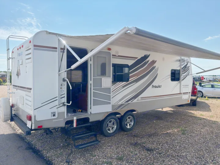 A white Prowler travel trailer, likely an rv rental, is set up on a gravel lot with its awning extended for outdoor use.. Fleetwood 230RKS 2009