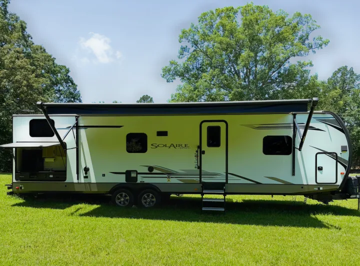 White Solaire travel trailer RV rental with outdoor kitchen, extended awning, and steps on green grass.. Palomino SolAire 320 TSBH 2022