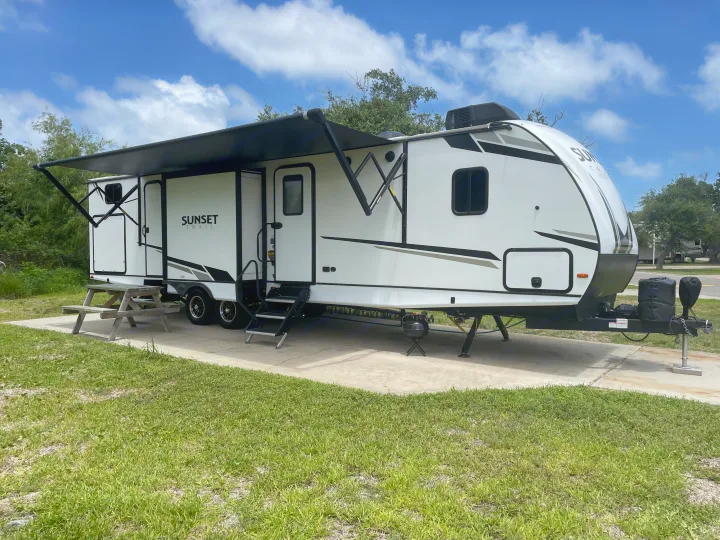 A white travel trailer with an extended black awning and steps, parked on a concrete pad at a campsite. Great RV rental option!. Crossroads RV Sunset Trail Super Lite 2023