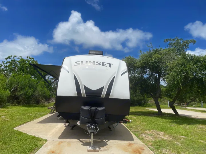 A 'Sunset Trail' RV rental is parked on a concrete pad at a sunny campsite, surrounded by green grass and trees.. Crossroads RV Sunset Trail Super Lite 2023
