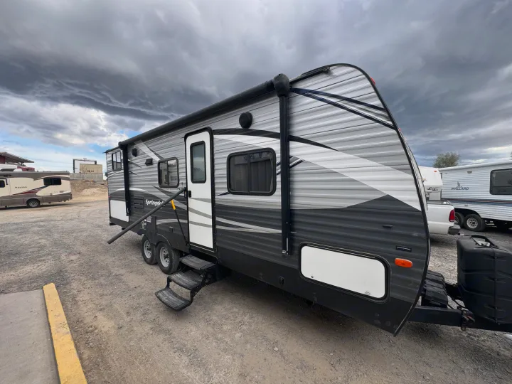 A modern grey travel trailer with a white door and awning in a gravel lot under cloudy skies. Perfect for an RV rental.. Keystone RV Springdale 2018