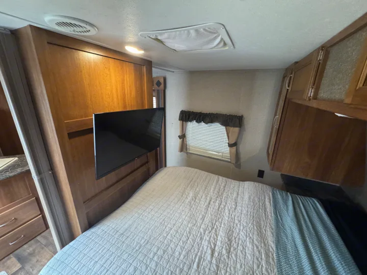 Cozy RV rental bedroom features a comfy bed, wall-mounted TV, window with blinds, and ample wood cabinets.. Keystone RV Springdale 2018