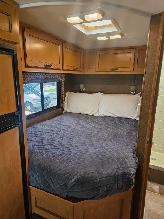 Cozy RV rental bedroom featuring a large bed with white pillows, overhead storage cabinets, a window, and a glimpse of the shower.. Thor Four Winds 2016