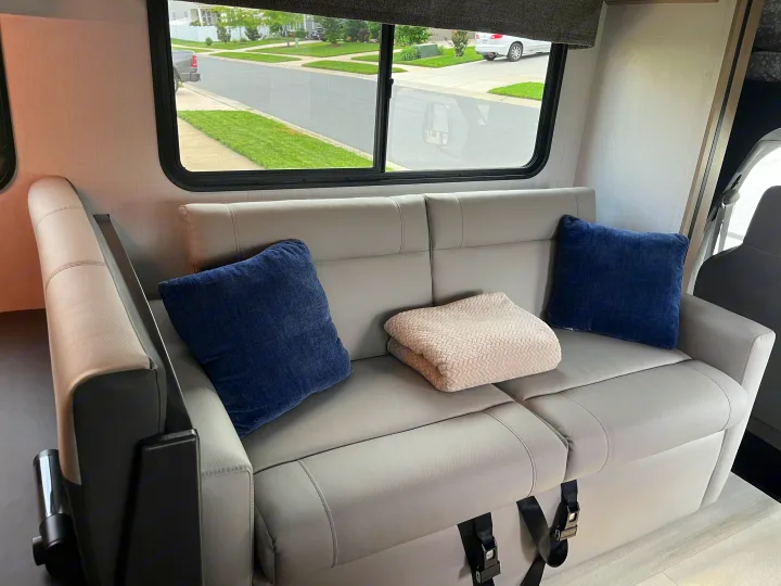 RV rental living area featuring a gray sofa, soft pillows, and a blanket, offering a view of a suburban street.. Forest River Forester 2024