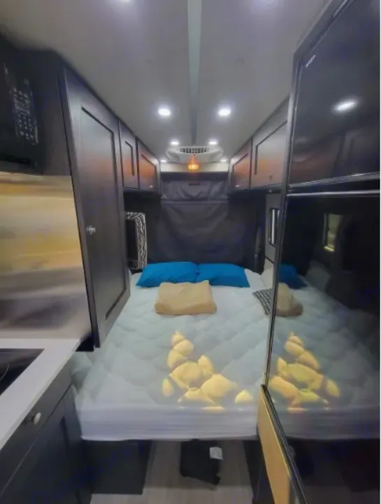 Inside a modern rv rental, showing a comfortable bed, overhead cabinets, and a compact kitchenette setup.. Roadtrek Roadtrek Class B 2021