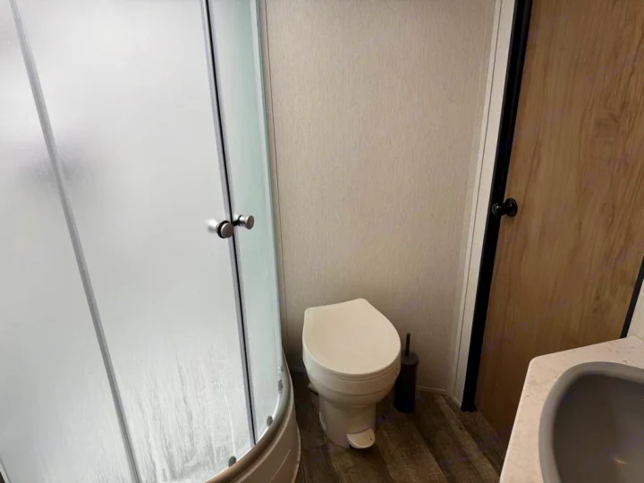 RV rental bathroom features a frosted glass shower, white toilet, and a wooden door. A compact, functional space with wood-look flooring.. Forest River Cherokee Grey Wolf 2023