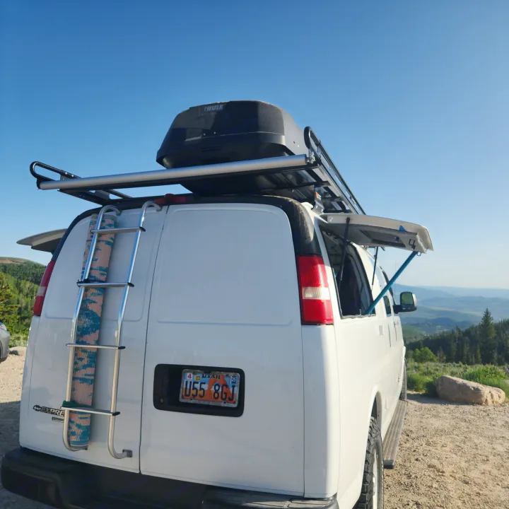 White van RV rental with roof box, ladder, and side awning, parked on a scenic overlook with mountains and blue sky.. Chevrolet Express 1500 AWD 2014