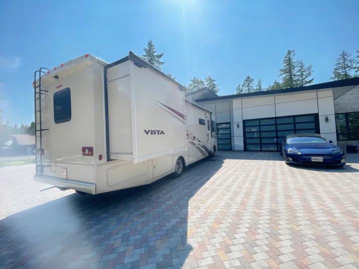 Spacious RV rental parked on a patterned driveway next to a modern house and blue Tesla under a sunny sky.. Winnebago Vista 2019