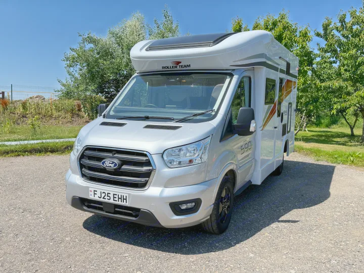 Silver Ford RV rental parked on gravel under a clear blue sky, surrounded by green trees. Ideal for travel.. Rollerteam Kronos Compact 590 2026