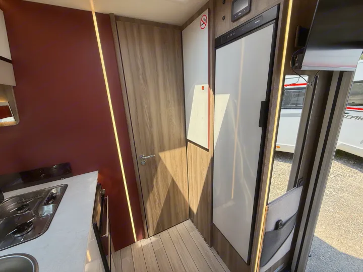 Step inside this modern rv rental interior: compact kitchen with sink/stove, integrated fridge, wood door, and a maroon accent wall with LED strip lighting.. Rollerteam Kronos Compact 590 2026