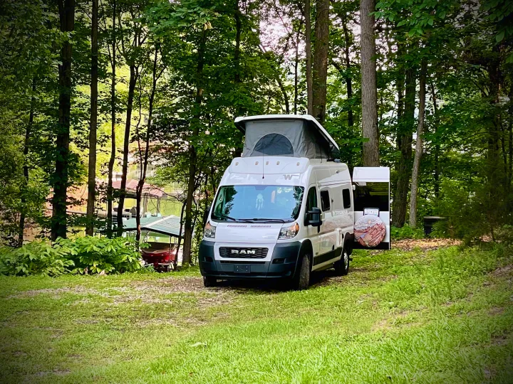 White Winnebago RV rental camper van parked at a wooded lake campsite with pop-top roof extended.. Dodge Winnebago 2021