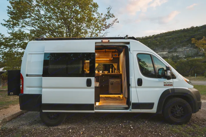Step inside this cozy Ram ProMaster RV rental, featuring a custom interior kitchen with warm lighting, parked amidst nature.. Dodge 1500 2020